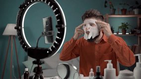 Male beauty blogger. Vlogger applying beauty sheet mask while recording video and giving tips for his beauty blog. Young bearded man testing new product. Cosmetic sheet face mask