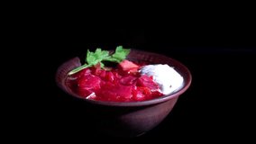 A plate of borscht with sour cream and herbs on a black background, illuminated by a bright side light. 
