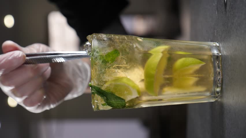Prepairing Fresh Mojito Drink on Bar. Slow motion. Vertical footage. Mojito cocktail in glass. Close up soda water with fresh slices lime, mint and ice is stirred with spoon. Carbonated drink. Summer | Shutterstock HD Video #1111229775