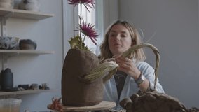 Embark on a journey of artistic discovery as a fearless female artist, amidst scattered materials and tools, passionately creates a unique bouquet of flowers and wild plants in a clay vase. This video