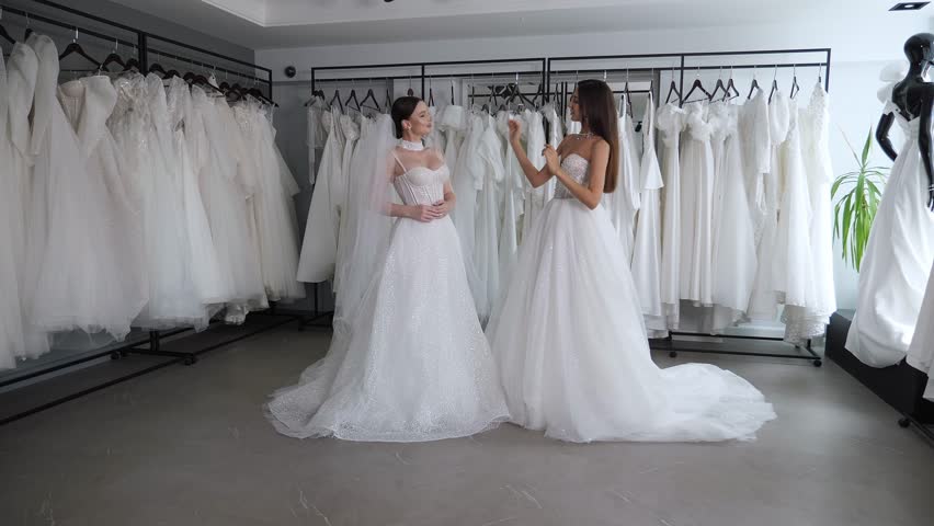 Two gorgeous women in wedding dresses in a wedding salon. Fitting wedding dresses in a modern showroom. Two friends try on wedding dresses and a veil together. Royalty-Free Stock Footage #1111232297
