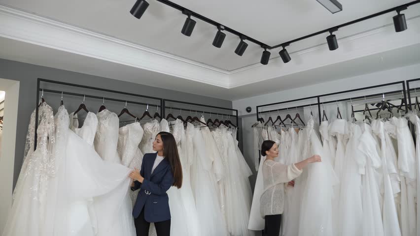 Two happy girlfriends in the salon are choosing a wedding dress, preparing for a wedding celebration. Two women in a bridal salon are looking at different wedding outfits. Royalty-Free Stock Footage #1111232303
