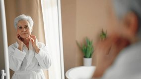 Senior wellbeing smiling woman in bathrobe with gray hair looking at mirror touching face worrying about aging process. Good looking old lady searching face wrinkles. Getting old age change concept. - Powered by Shutterstock - Get 15% off with code: PIKWIZARD15