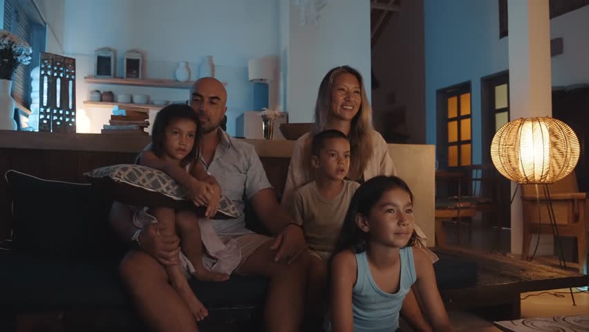 Family of Happy Woman and Man with Young Child Watching TV and Sitting on Sofa. People on Couch Inside at Home Together Watch Television. Pretty Mixed Race Siblings Looking at News on Digital Display