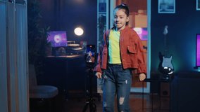 Kid doing viral dance choreography in living room with 3D rendered animations on computer screens in background. Child dancing in neon lit home studio interior, producing content with cellphone