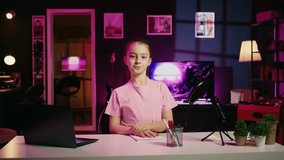 Young child filming in living room used as professional studio for recording vlogs for online channel. Smiling kid sitting down at desk in pink neon lit apartment discussing topics with young audience