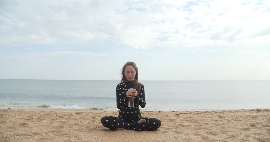 A young woman in a black skinny suit with white polka dots sits on a sandy beach in the lotus position and drinks water from a transparent plastic bottle against the backdrop of the beach and horizon