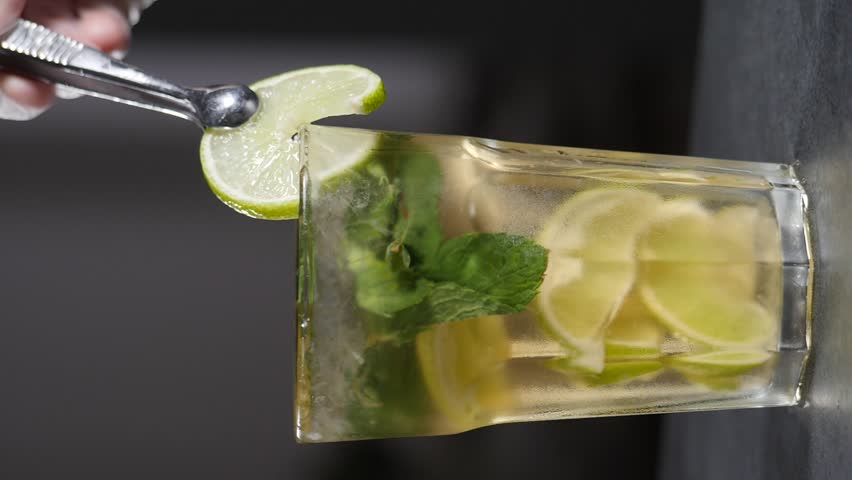 Preparing Lemonade presentation serving cocktail. Putting lemon slice on glass cup with ice. Slow motion. Vertical footage. Cocktail mojito with fresh lime. Detox cold tonic water with sunny lemon | Shutterstock HD Video #1111244107