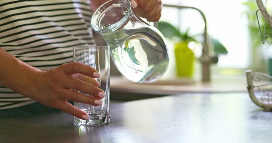 Close up of woman pouring fresh clean water into glass from jug at home