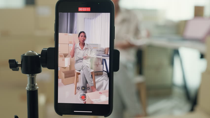 Close up youth Asia lady owner small business online shop sit front of phone look at camera live streaming talk to customer order promotion with full carton box at home. Shipping and delivery concept. | Shutterstock HD Video #1111255051