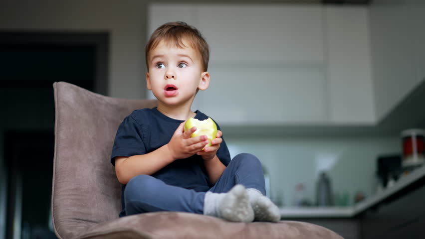 Beautiful little kid sitting on the soft chair in the kitchen. Cute child eating tasty big apple. Low angle view. Blurred backdrop. Royalty-Free Stock Footage #1111255137