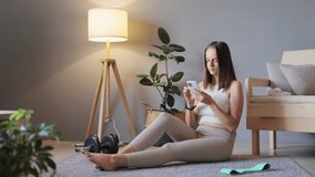 Smartphone for workouts. Mobile exercise tracking. Home workout and wellness. Slim woman wearing beige sportswear sitting on floor in living room in home interior.
