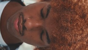 Vertical video, Closeup, guy with an African hairstyle dressed in blue shirt raises his head, opens his eyes and smiles looking at the camera