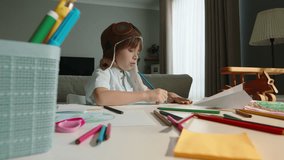 Boy in pilot's hat is drawing, wooden plane on table. World of child artist, pencil becomes magic wand, turning thoughts and dreams into tangible creations. In dreams of becoming airplane pilot