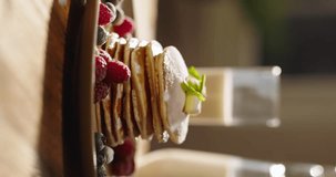 Side view of pancakes adorned with juicy berries, syrup, and sugar powder, kitchen. Leisurely cozy morning, essence of lifestyle, food, cooking, and domestic life. Vertical video, no people, cinematic