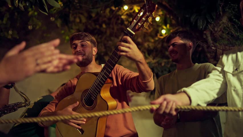 Handsome young guy playing Spanish guitar while his friends clap at night. Slow motion  Royalty-Free Stock Footage #1111258217