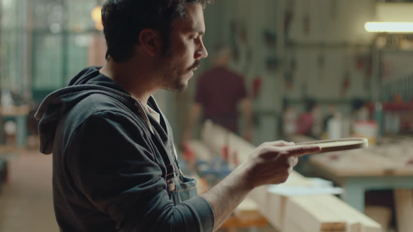 Young male woodworker standing in carpentry workshop, blowing sawdust off the wooden tray and checking its smoothness after polishing. Side view, medium shot