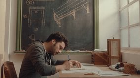 Young furniture master drawing sketches at desk near the window, working on project design in a woodworking workshop. Side view, copy space - Powered by Shutterstock - Get 15% off with code: PIKWIZARD15