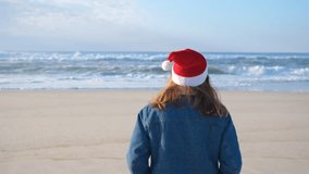 A girl or young woman in a red Santa Claus hat walks slowly along the shore of the Atlantic Ocean. Christmas or New Year holiday concept. 4K resolution video.