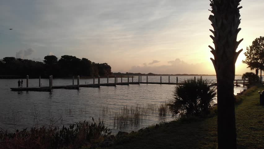 Sun shining bird passing by aerial drone wide shot of marina boat dock with lighthouse at the bay palm trees