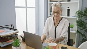Elegant grey-haired senior woman worker smiling while successfully managing business work; having an important video call at the office indoors on her laptop.