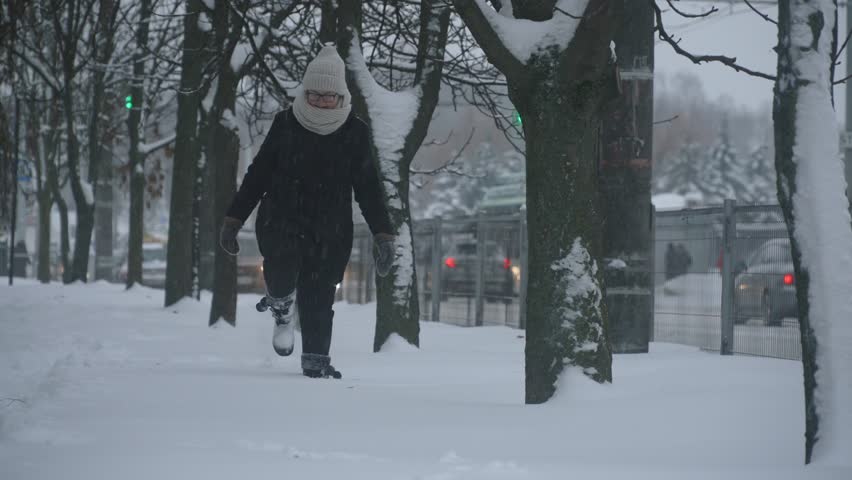 Woman walks with difficulty through deep snow on uncleaned sidewalk near snow covered trees in city, handheld shot. Cars drive next to person in difficult weather conditions on winter day.
