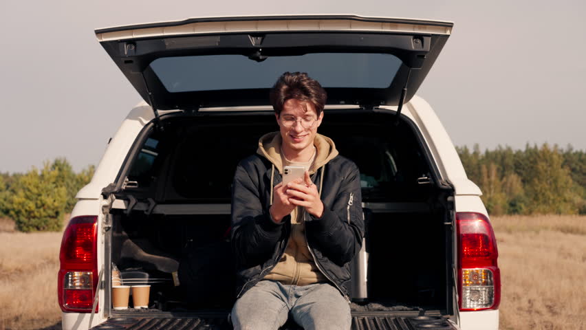 A young guy sits in the open trunk of a car with phone and studies the area during a stop in a meadow