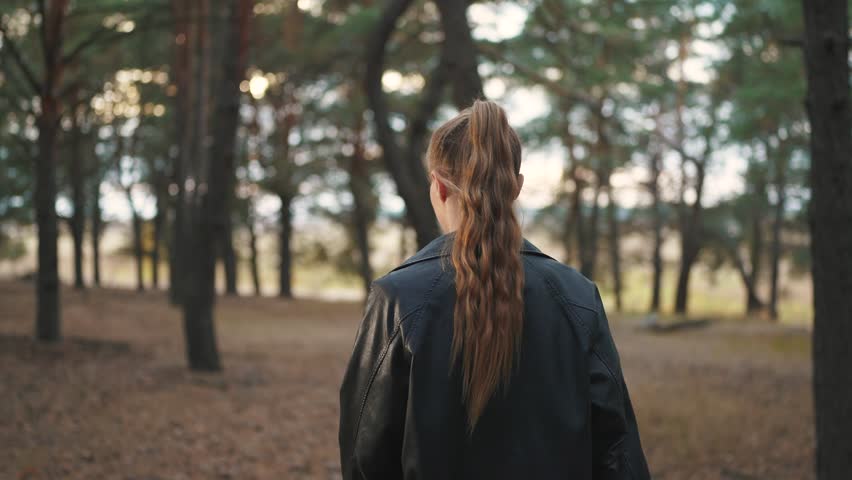 girl walking through the forest. nature dream concept. a girl with a ponytail on her head and wearing a leather jacket walks through the autumn evening coniferous forest along the road