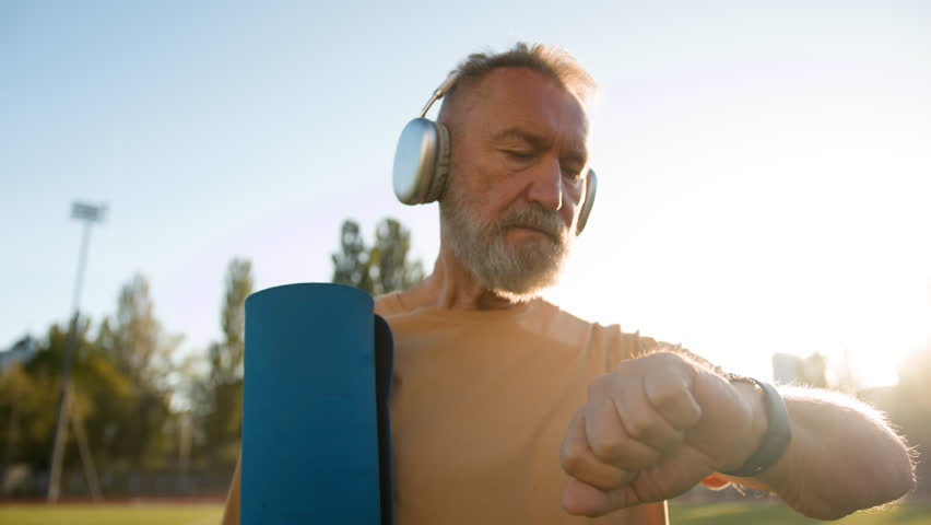Elderly Caucasian man in headphones looking at wristwatch sport time morning stadium exercise athlete training mat physical activity lifestyle health wellbeing city outside motivated fitness equipment Royalty-Free Stock Footage #1111296977