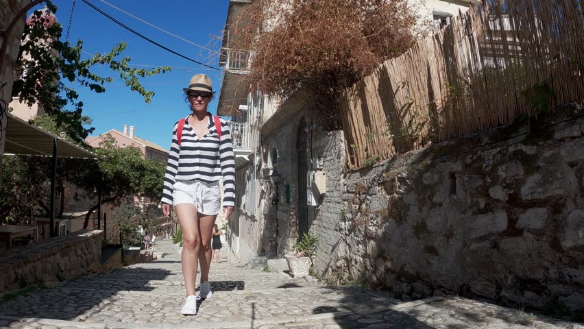 Woman tourist in a hat walks on the street stairs of the ancient Greek city. Corfu Island in Greece, slow motion shot