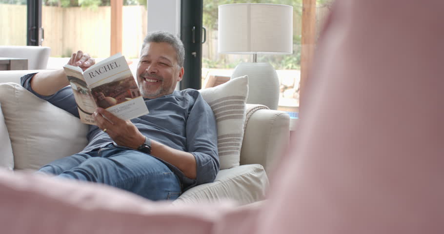 Happy senior biracial man relaxing on couch reading book in living room, copy space, slow motion. Reading, relaxation, hobbies, retirement, domestic life and senior lifestyle, unaltered.