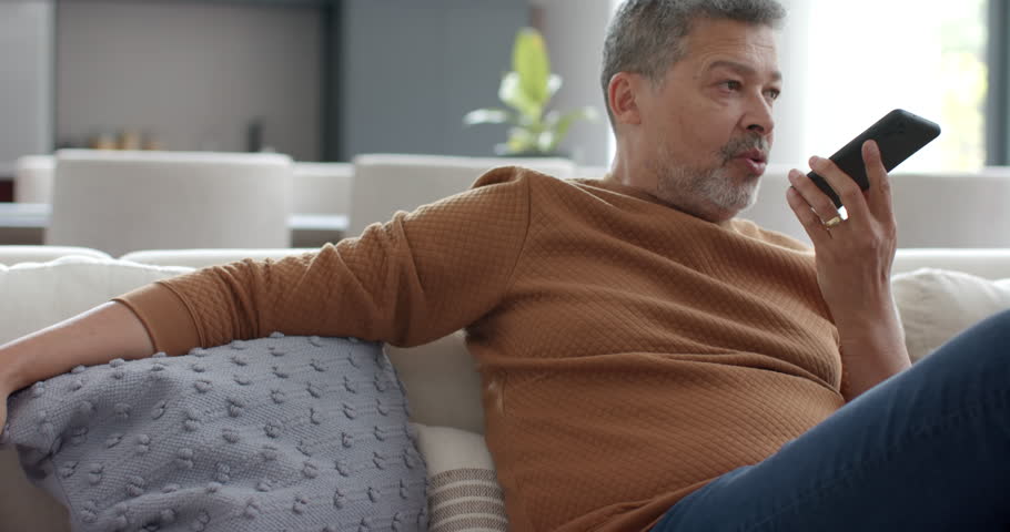 Senior biracial man talking on smartphone sitting on couch in living room, slow motion. Communication, retirement, domestic life and senior lifestyle, unaltered.
