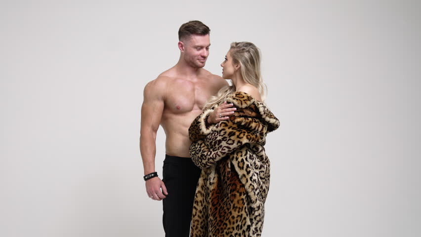 Man with athletic body stands near an attractive woman wearing fur-coat. Passionate couple in studio together. White backdrop. Royalty-Free Stock Footage #1111305431