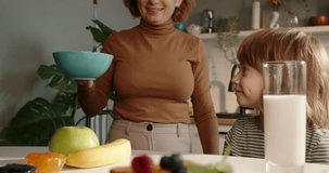 Mother places bowl of breakfast cereal for son as he sits at the table in bright modern kitchen. Promotional video, cinematic in slow motion. Happy family concept. Balanced healthy breakfast nutrition