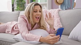 Young blonde woman having video call lying on sofa at home