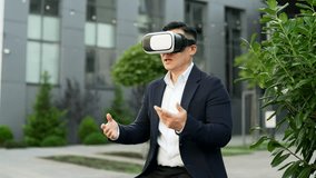 Asian businessman in formal suit using VR glasses for video call and chatting online in virtual reality simulator while sitting on a bench on street near office building. Online conference remotely