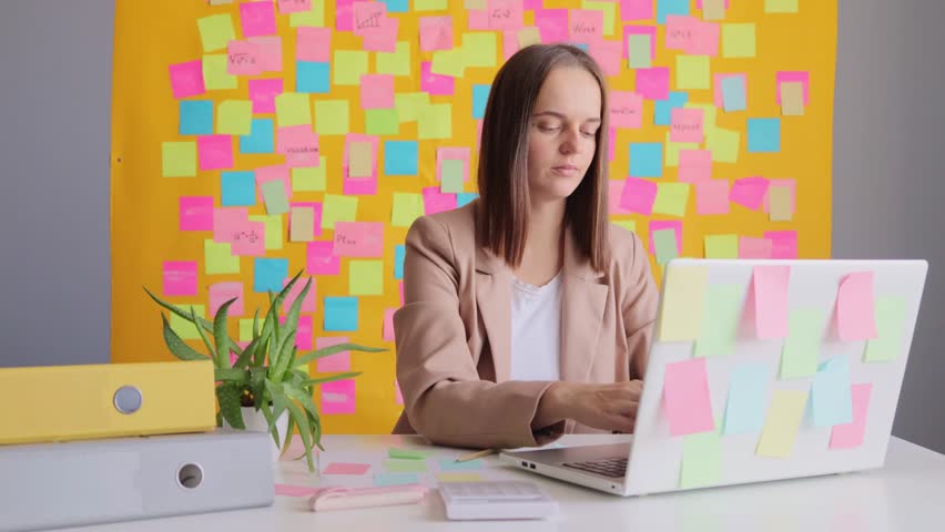 Sad exhausted beautiful business woman wearing beige jacket sitting in office using laptop computer against yellow wall with multicolored stickers having lots of office work.