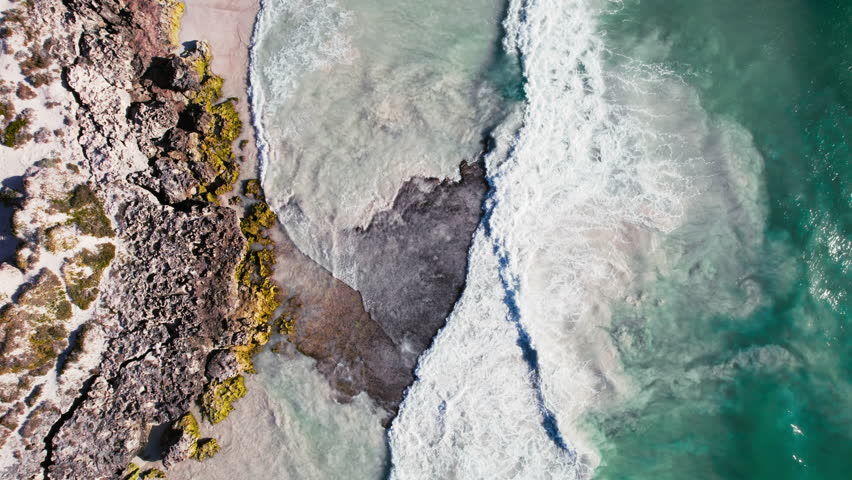 Top down view on turquoise ocean waves crashing and foaming around majestic rocks and beach with white sand. Drone shot of powerful ocean water on epic coastline, incredible nature concept. | Shutterstock HD Video #1111354093