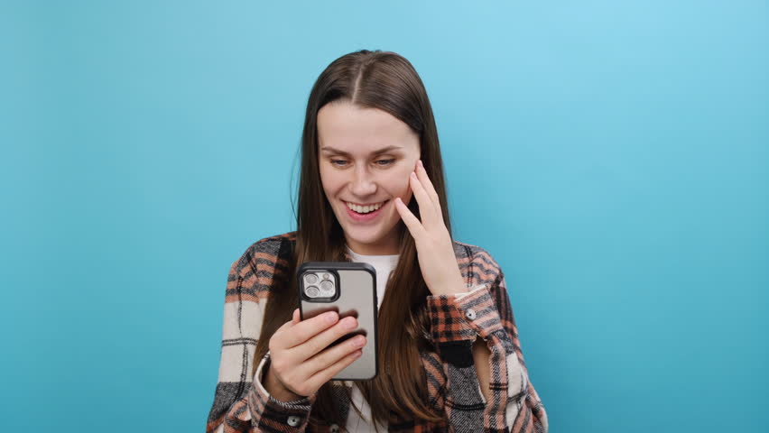 Excited surprised joyful young caucasian woman wear casual shirt use pointing finger on mobile cell phone just found out great big win news, posing isolated over plain pastel blue background studio