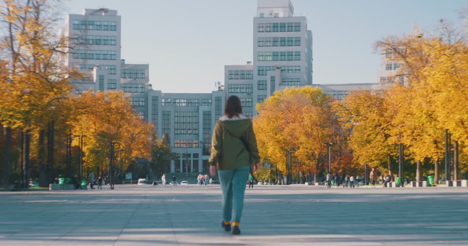 A young girl walks forward along a wide city street against the background of a building, the camera rises up to the building and the sky