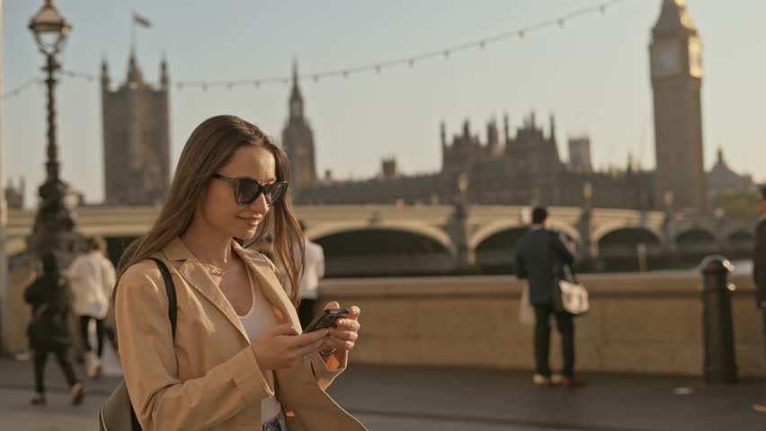 Stylish young beautiful woman smiling and using smartphone while walking with London Big Ben Palace of Westminster background view Royalty-Free Stock Footage #1111373181