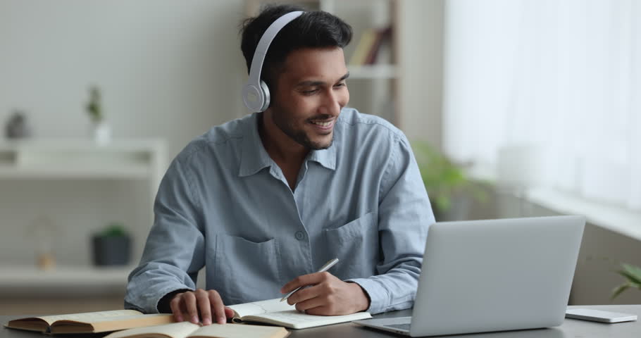 Focused serious Indian student guy in headphones watching online learning conference on laptop, talking on video call to teacher, studying, writing notes, speaking Royalty-Free Stock Footage #1111376725