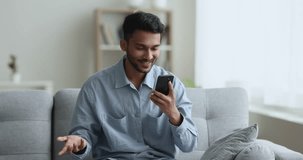 Cheerful young Indian guy talking on video conference call on smartphone, holding cellphone, speaking, smiling, enjoying online conversation, looking at screen, sitting on soft home couch