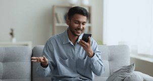 Cheerful young Indian guy talking on video conference call on smartphone, holding cellphone, speaking, smiling, enjoying online conversation, looking at screen, sitting on soft home couch