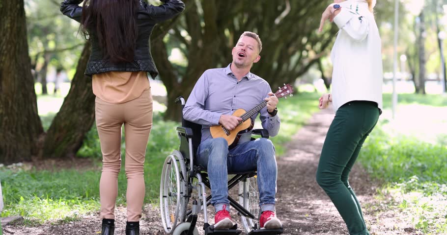 Young disabled man in wheelchair playing ukulele in front of dancing women in park 4k movie. Full life of people with disabilities concept Royalty-Free Stock Footage #1111384671