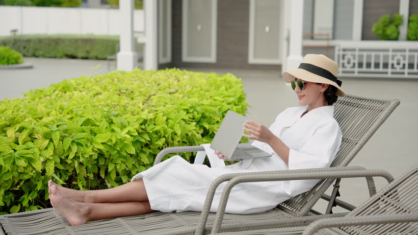 Beautiful Hispanic girl wearing a white coat After leaving the pool Soaking up the sun happily on a chair by the pool Wear a black swimsuit Wear a hat and sunglasses. On a clear summer day