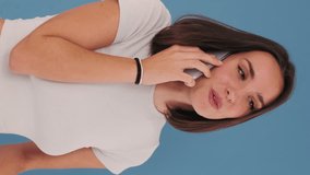 Vertical video, Young woman dressed in white top surprised while talking on mobile phone, , happy news, isolated on blue background in studio