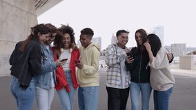 Young group of happy people laughing together using mobile phone outdoors. Smiling multiracial student friends having fun watching funny social media videos on smartphone app. Youth community concept