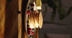 Elegant process of decorating homemade pancakes with berries. Unknown woman places raspberries sprig of mint on stack of pancakes. Magic of morning, cozy kitchen. Cinematic vertical video, advertising