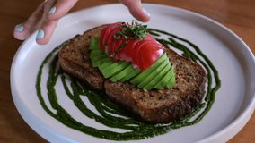 Young woman takes avocado toast with tomatoes from plate. Slow motion video of decorated plate with sliced avocado, tomatoes on toast and sauce on plate. Concept of eating healthy avocado toast